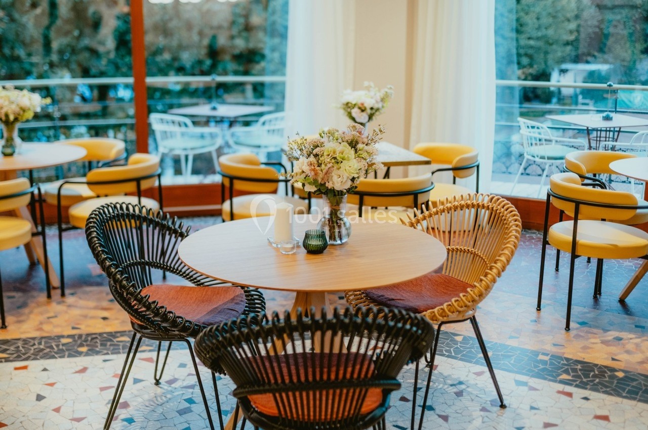 Salle lumineuse avec des tables rondes, chaises en rotin et un décor floral, donnant sur une terrasse vitrée.