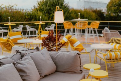 Terrasse ensoleillée avec tables rondes blanches, chaises et coussins jaunes, vue sur des toits et des arbres.