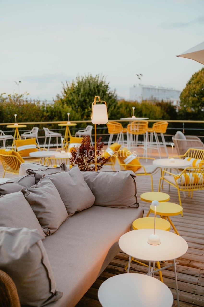 Terrasse aménagée avec des meubles jaunes et gris, comprenant des tables, des chaises et un canapé, entourée de verdure.