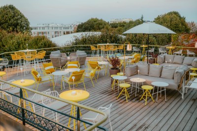 Terrasse ensoleillée avec tables rondes blanches, chaises et coussins jaunes, vue sur des toits et des arbres.
