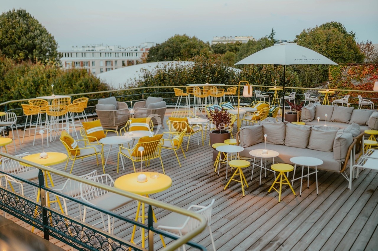 Terrasse en bois aménagée avec des tables et chaises jaunes, des canapés gris et un parasol, entourée de verdure.