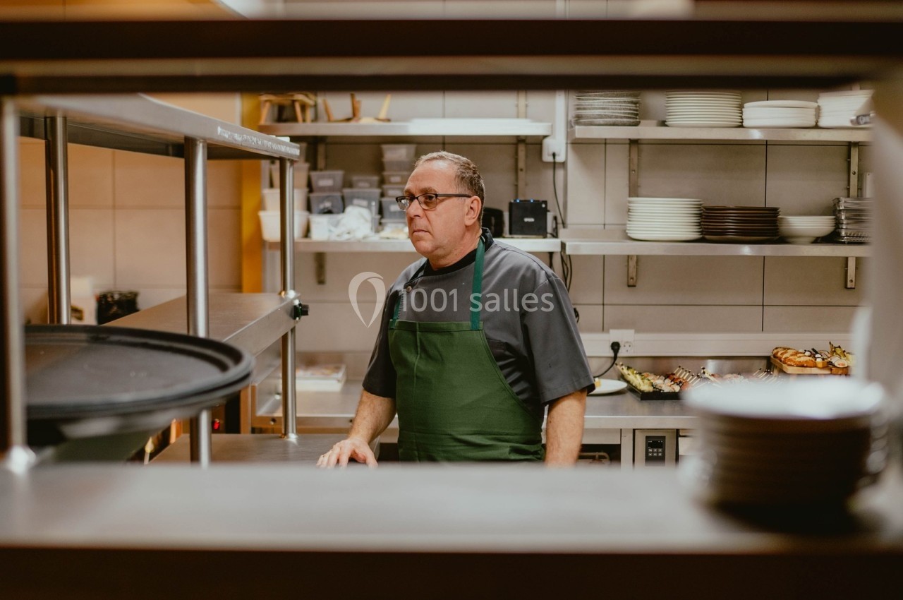 Un cuisinier en tablier vert se tient dans une cuisine professionnelle entourée d'étagères et d'ustensiles.