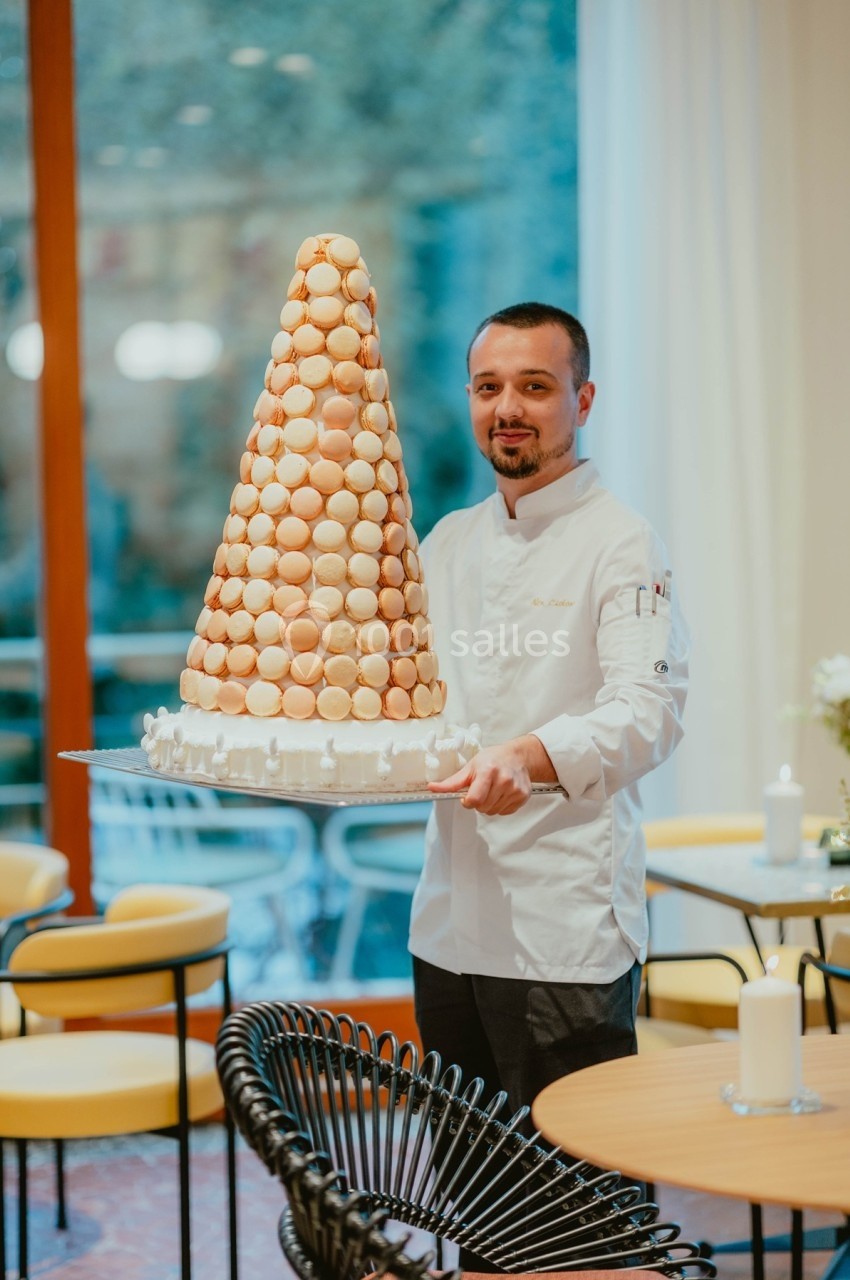 Un pâtissier en tenue blanche présente une pièce montée de macarons dans un cadre de restaurant lumineux.