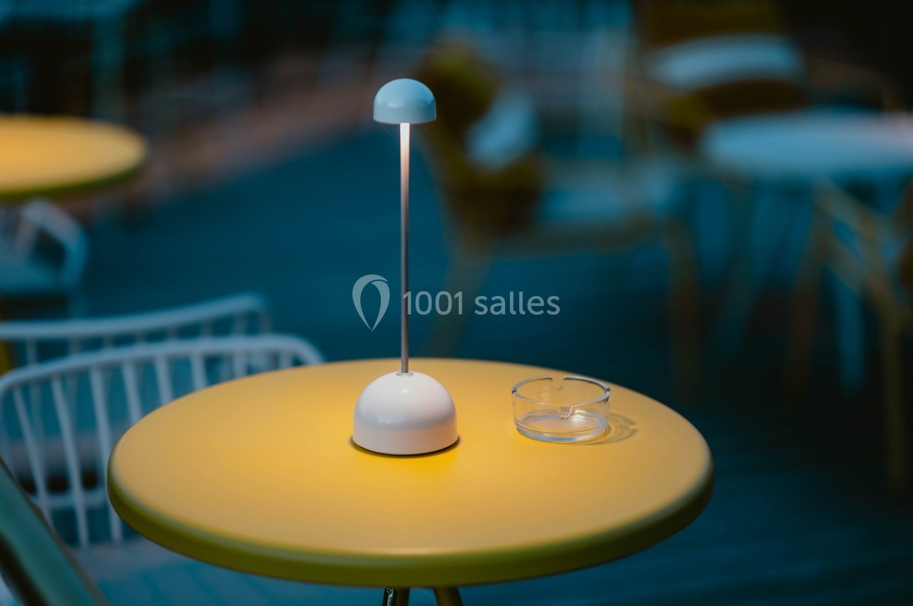 Lampe de table allumée sur une table jaune avec un cendrier en verre, en extérieur, ambiance nocturne floutée.
