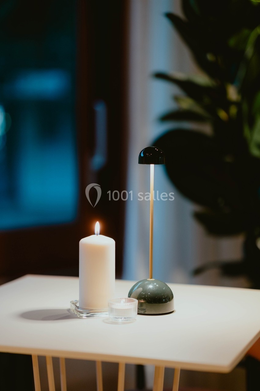 Bougie allumée et petite lampe moderne sur une table blanche, avec une plante et une fenêtre en arrière-plan.