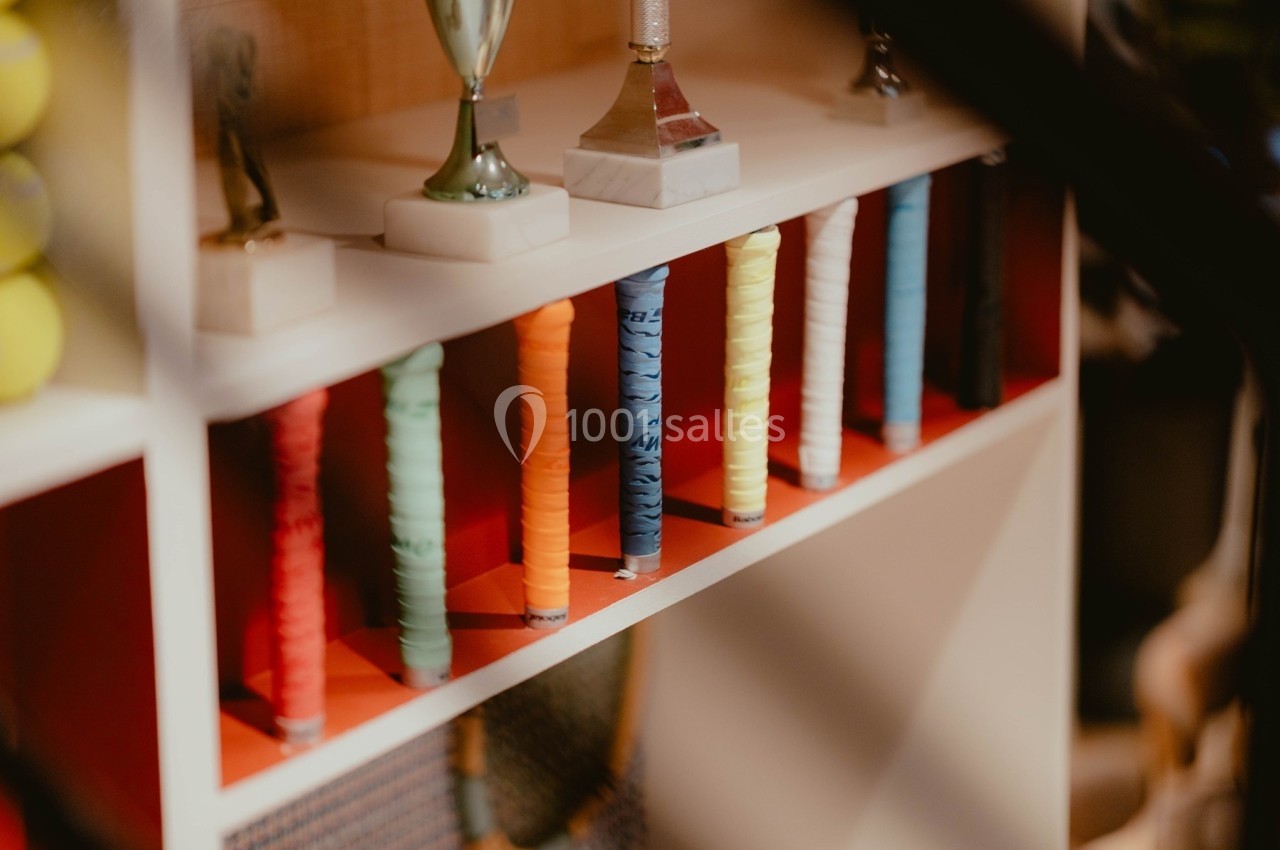 Raquettes de tennis avec poignées colorées alignées sur une étagère, entourées de trophées et d'autres objets.