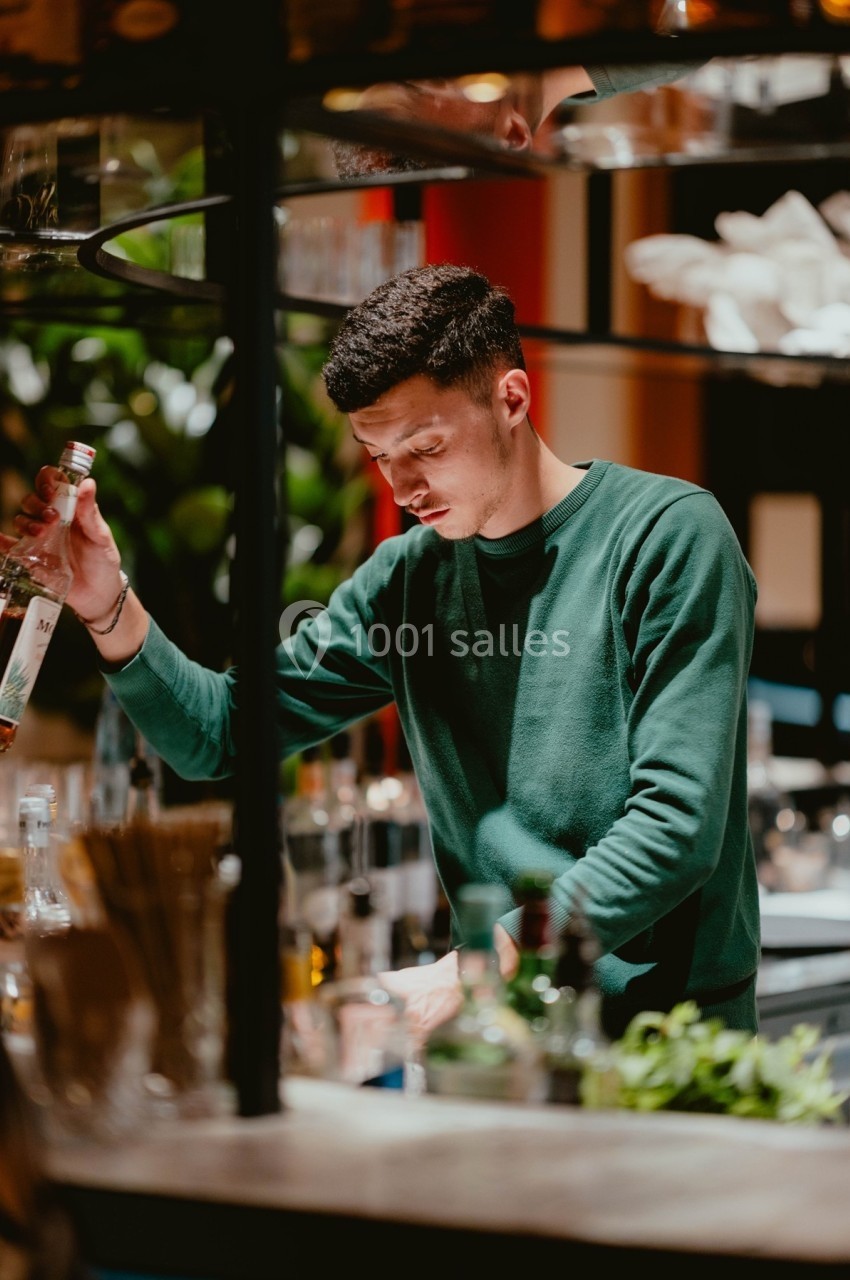 Un barman en pull vert prépare une boisson derrière un comptoir, entouré de bouteilles et d'ustensiles.