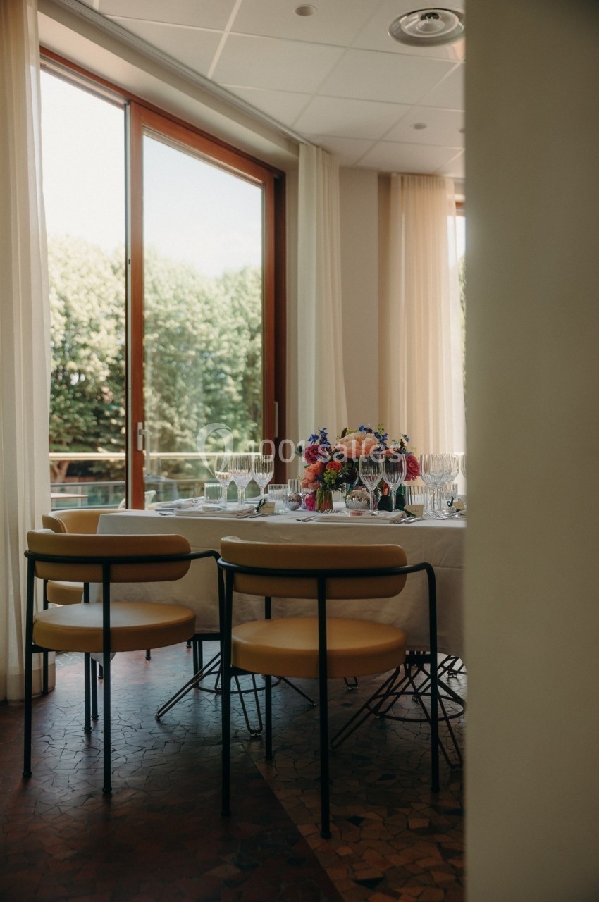 Salle lumineuse avec une table dressée, décorée de fleurs colorées, près de grandes baies vitrées donnant sur des arbres.