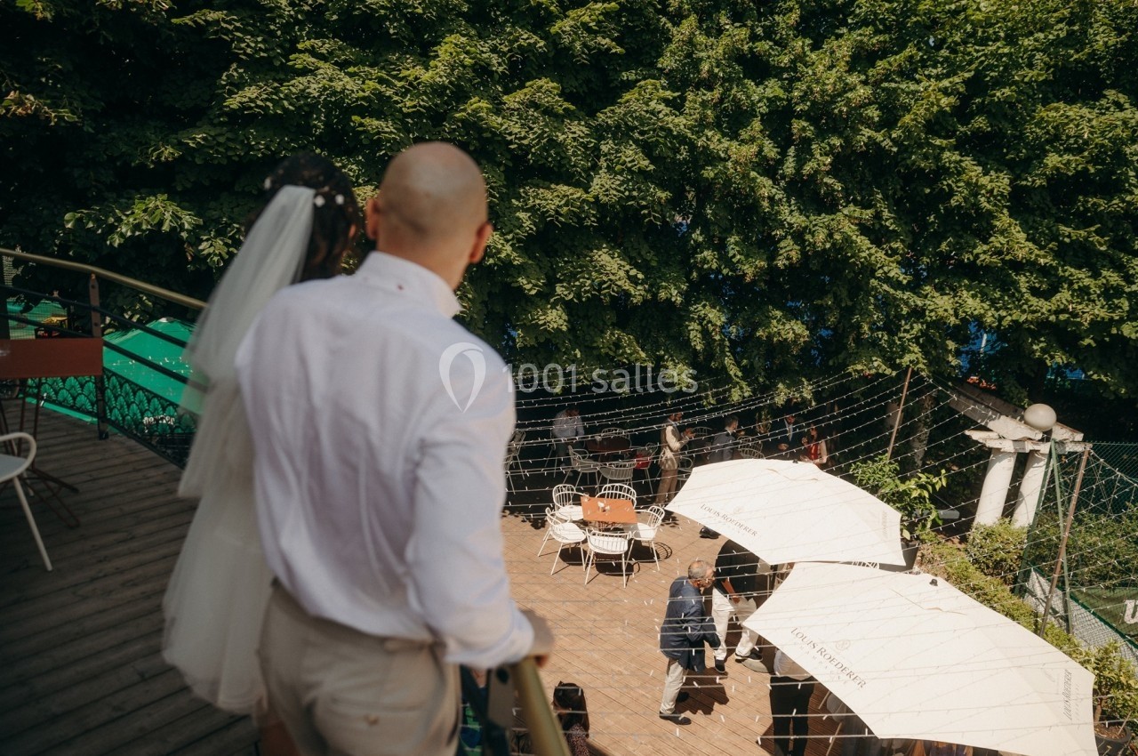 Un couple descend un escalier vers une terrasse extérieure avec des tables, des parasols et des arbres en arrière-plan.