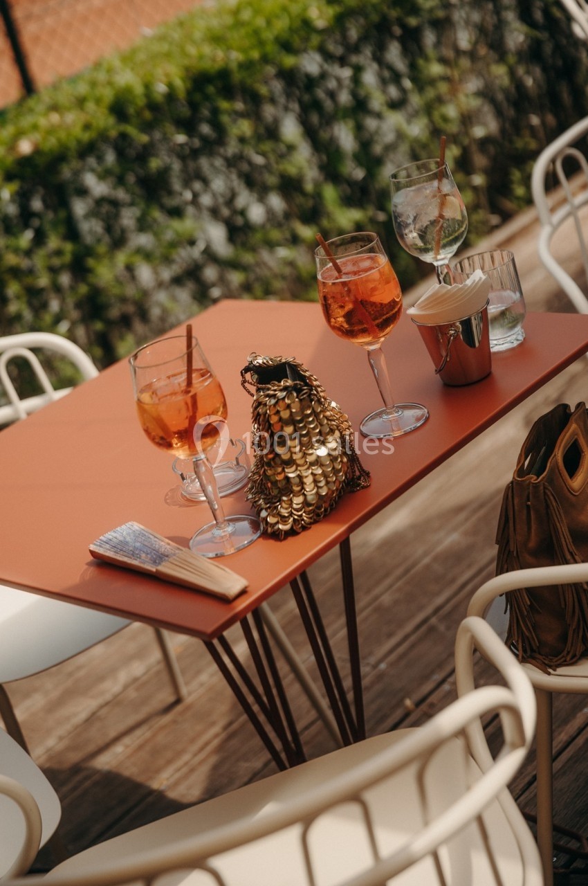 Verres de cocktail orange sur une table en terrasse, accompagnés d'un éventail et d'un sac à paillettes dorées.