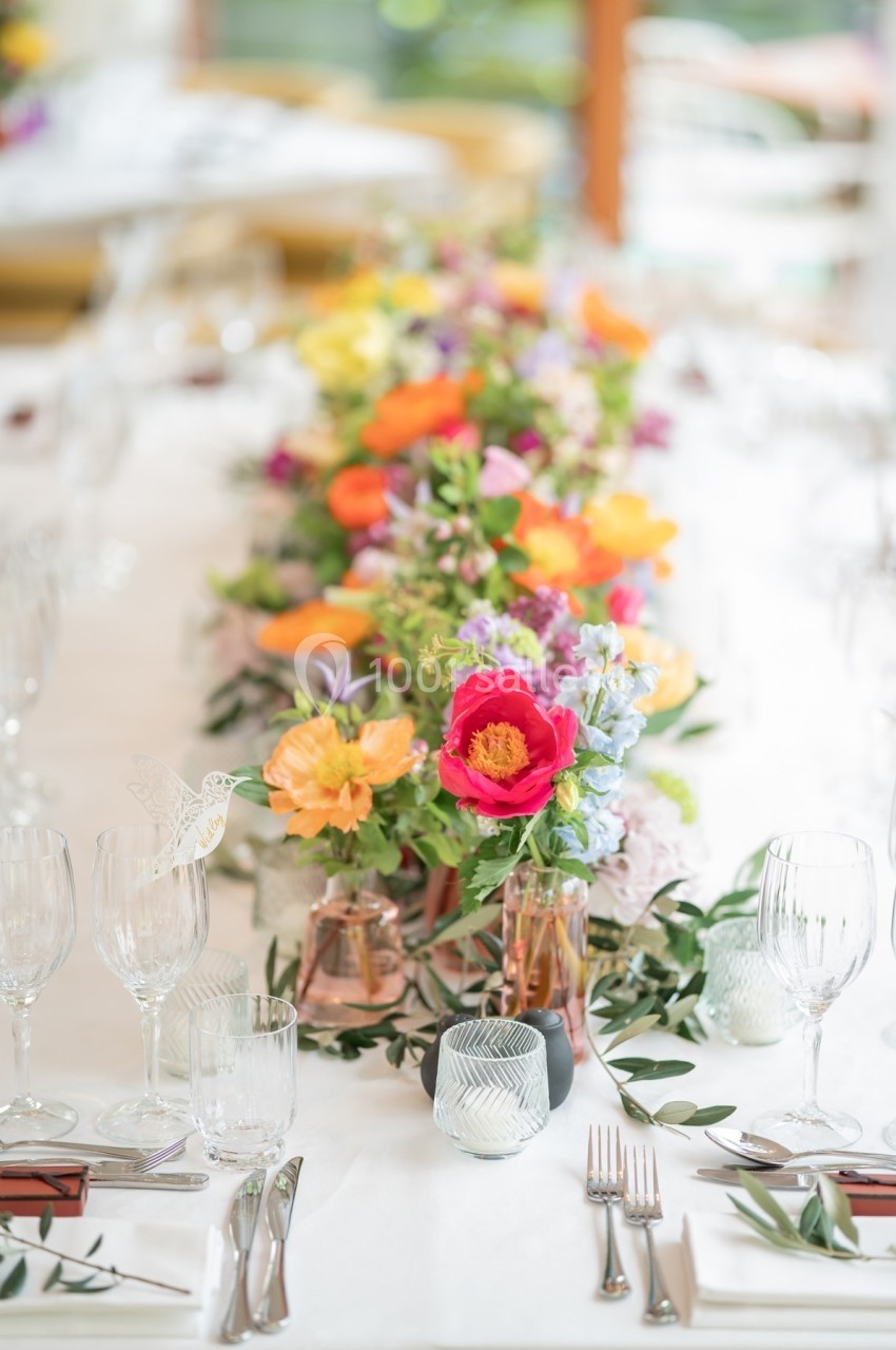 Table décorée avec un chemin de fleurs colorées, entourée de verres, couverts et bougies dans un cadre lumineux.