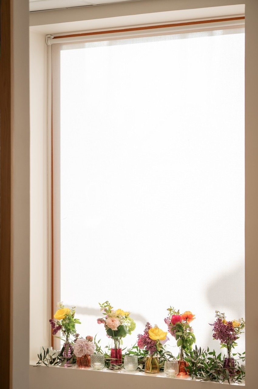 Fleurs colorées disposées dans des vases en verre sur un rebord de fenêtre avec un store blanc en arrière-plan.