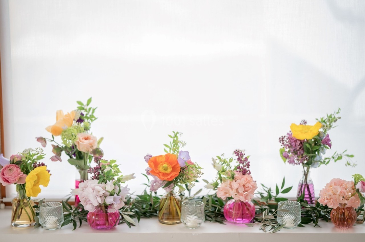 Vases colorés contenant des bouquets de fleurs variées disposés sur une surface blanche devant une fenêtre lumineuse.
