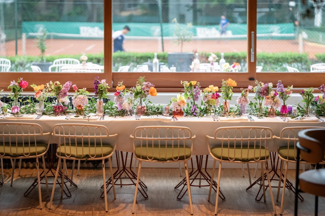 Table décorée avec des fleurs colorées, disposée pour un repas, avec vue sur un court de tennis en arrière-plan.