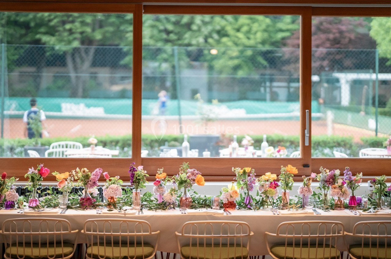 Table décorée avec des fleurs colorées devant une baie vitrée donnant sur un court de tennis en arrière-plan.