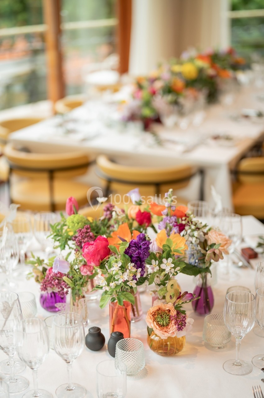 Tables élégamment dressées avec des bouquets de fleurs colorées, des verres et de la vaisselle dans une salle lumineuse.