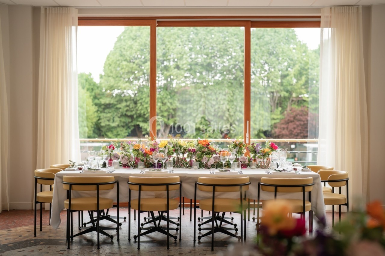 Table décorée avec des fleurs colorées, entourée de chaises, devant une grande baie vitrée donnant sur un jardin verdoyant.