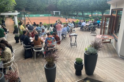 Terrasse ensoleillée avec tables rondes blanches, chaises et coussins jaunes, vue sur des toits et des arbres.