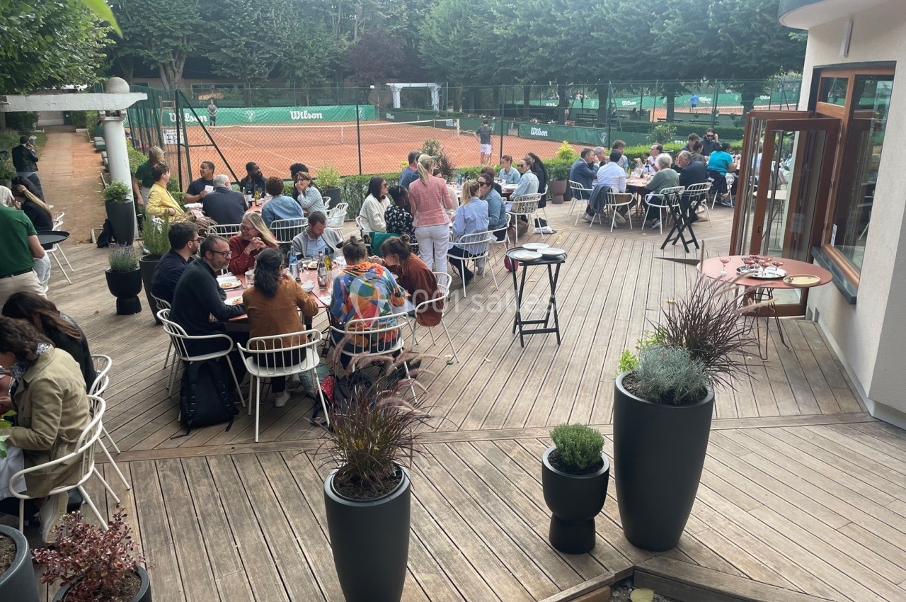 Personnes assises à des tables en terrasse en bois près d'un court de tennis, entourées de végétation.