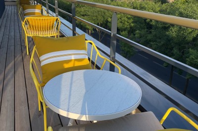 Terrasse ensoleillée avec tables rondes blanches, chaises et coussins jaunes, vue sur des toits et des arbres.
