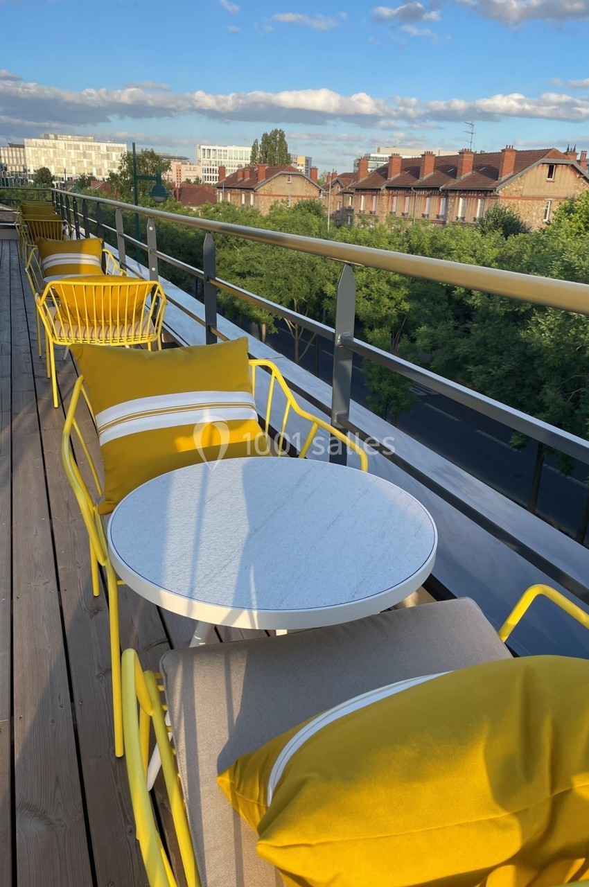 Terrasse ensoleillée avec tables rondes blanches, chaises et coussins jaunes, vue sur des toits et des arbres.