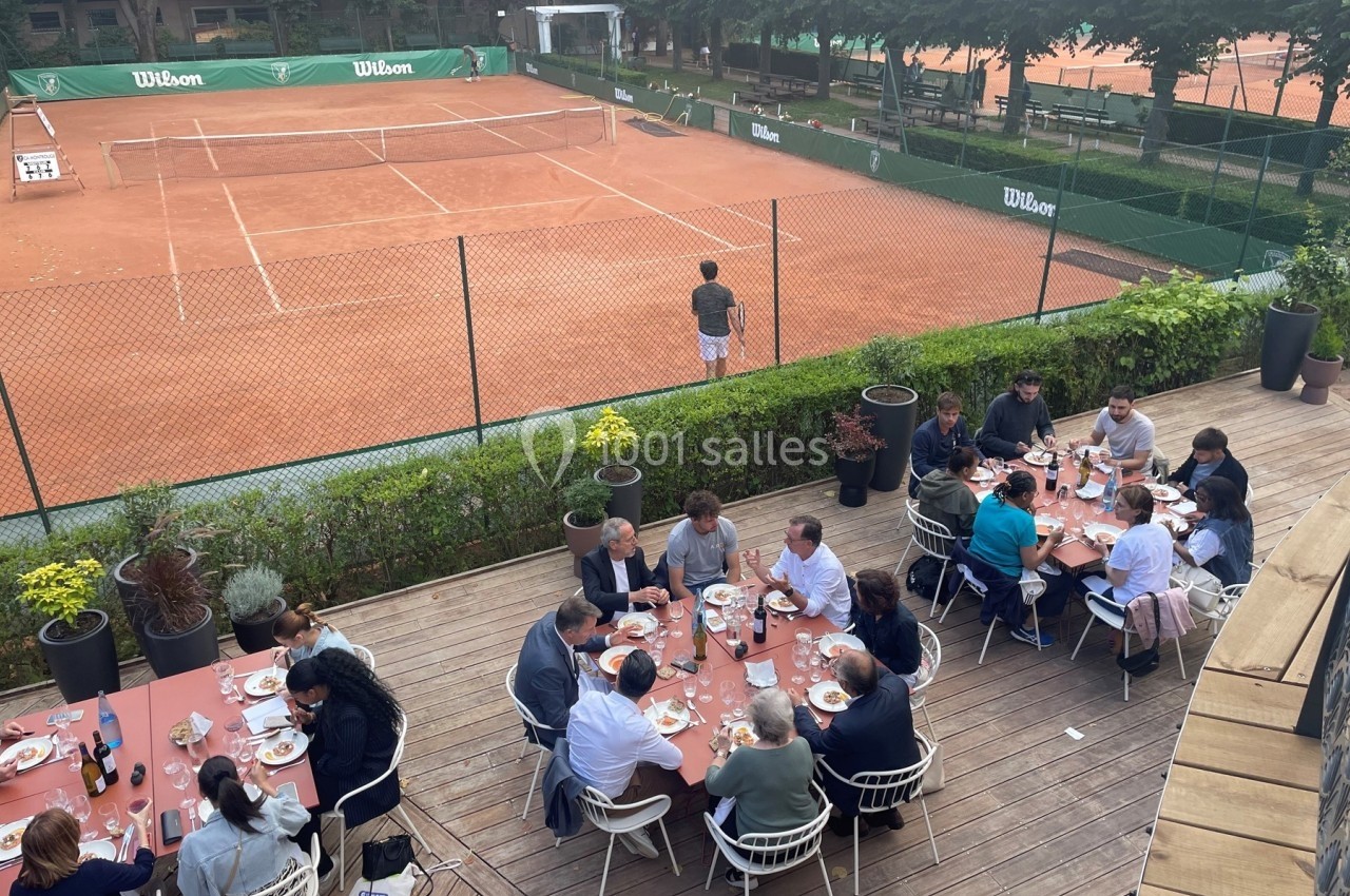 Des personnes dînent sur une terrasse en bois surplombant des courts de tennis en terre battue.