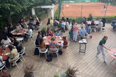 Terrasse ensoleillée avec tables rondes blanches, chaises et coussins jaunes, vue sur des toits et des arbres.