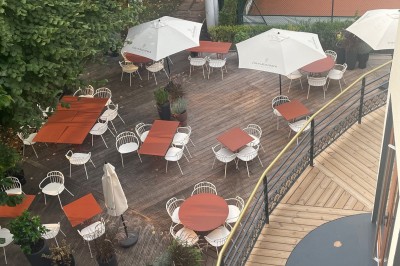 Terrasse ensoleillée avec tables rondes blanches, chaises et coussins jaunes, vue sur des toits et des arbres.