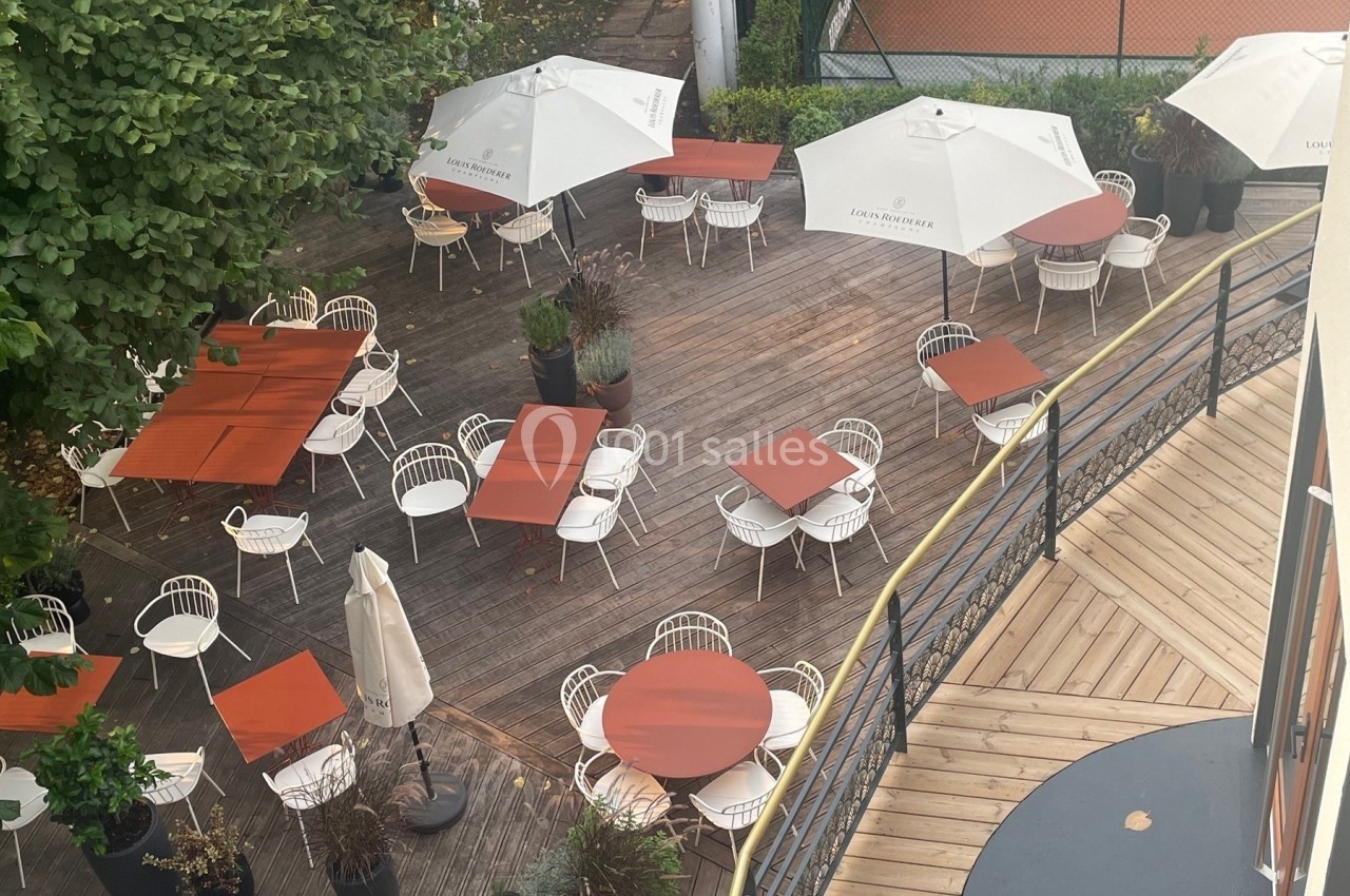 Terrasse en bois avec tables et chaises blanches, parasols ouverts, entourée de verdure et d'une clôture.