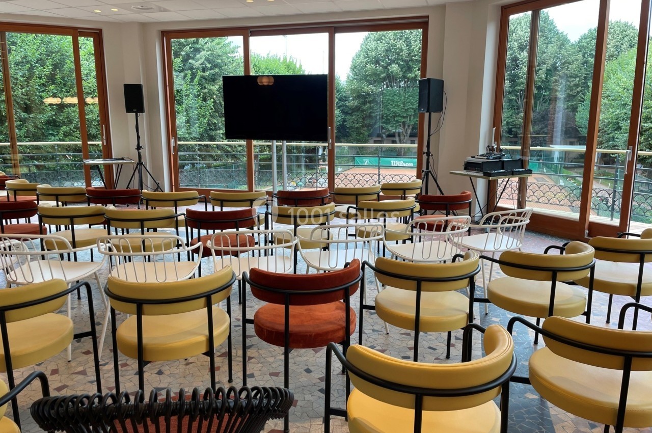 Salle de conférence lumineuse avec des chaises colorées, un écran noir et une vue sur un jardin à travers de grandes…