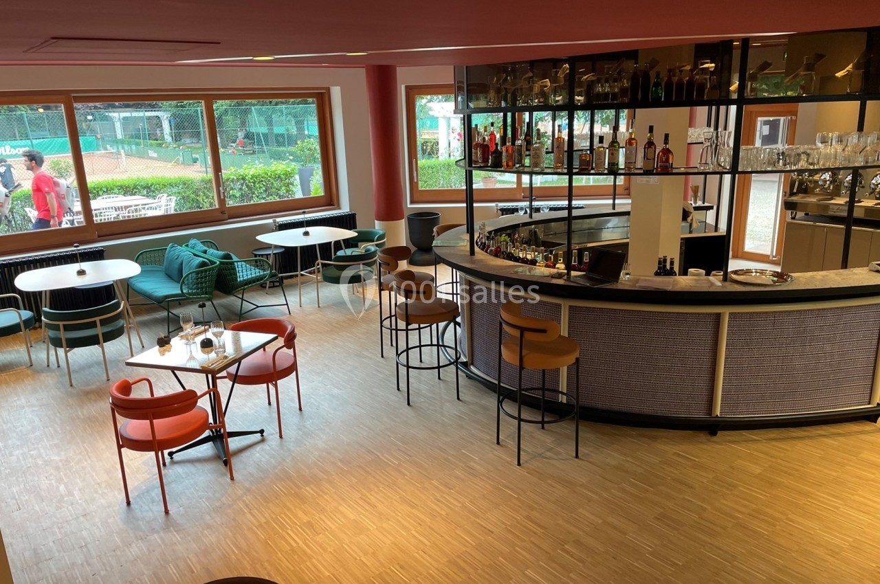 Vue d'un bar moderne avec comptoir arrondi, tables et chaises colorées, donnant sur des courts de tennis extérieurs.