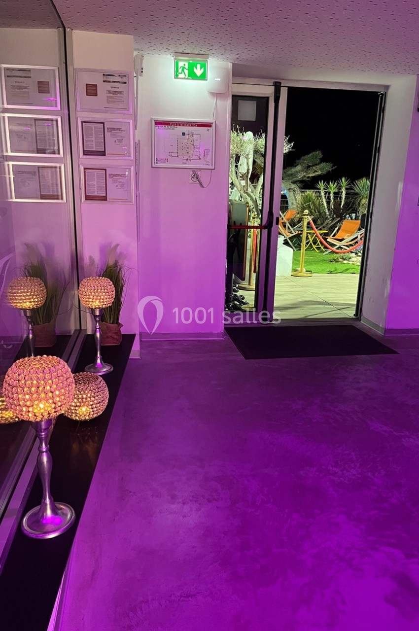 Entrée d’un bâtiment avec éclairage violet, porte ouverte sur une terrasse illuminée et décorée de plantes.