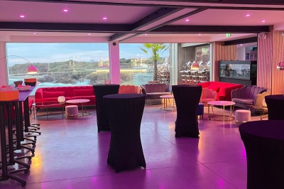 Des personnes assises à une table en terrasse face à la mer au coucher du soleil, avec des plantes au premier plan.