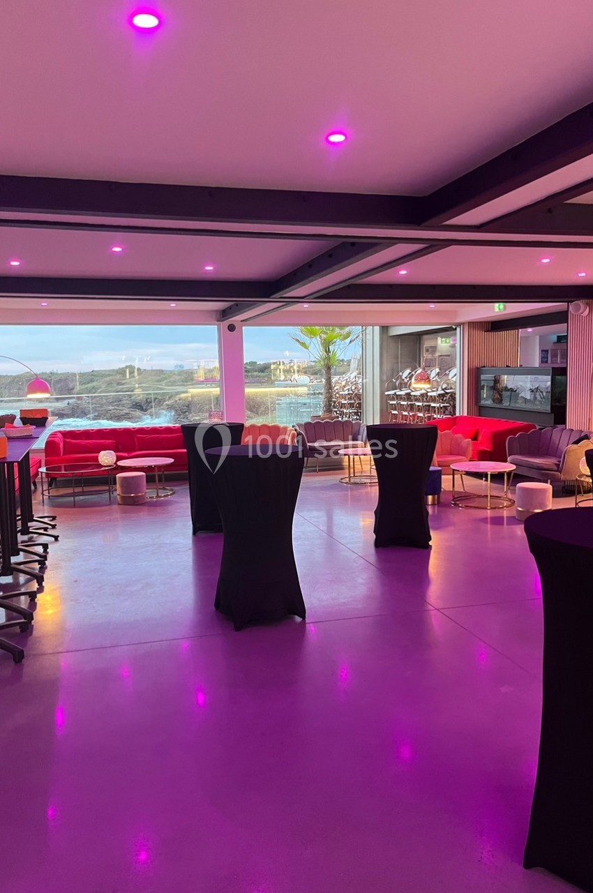 Salle moderne avec baies vitrées, mobilier coloré et tables hautes, éclairée par des lumières roses.