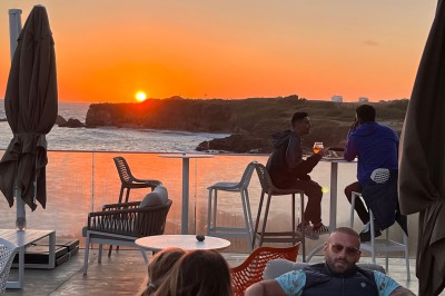 Des personnes assises à une table en terrasse face à la mer au coucher du soleil, avec des plantes au premier plan.