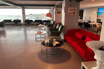 Des personnes assises à une table en terrasse face à la mer au coucher du soleil, avec des plantes au premier plan.