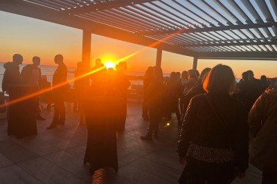 Des personnes assises à une table en terrasse face à la mer au coucher du soleil, avec des plantes au premier plan.