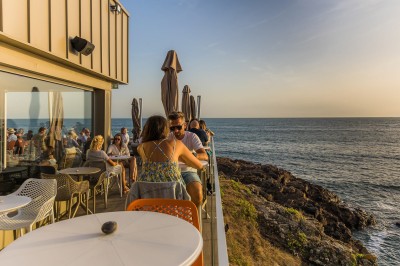 Des personnes assises à une table en terrasse face à la mer au coucher du soleil, avec des plantes au premier plan.