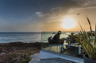 Des personnes assises à une table en terrasse face à la mer au coucher du soleil, avec des plantes au premier plan.