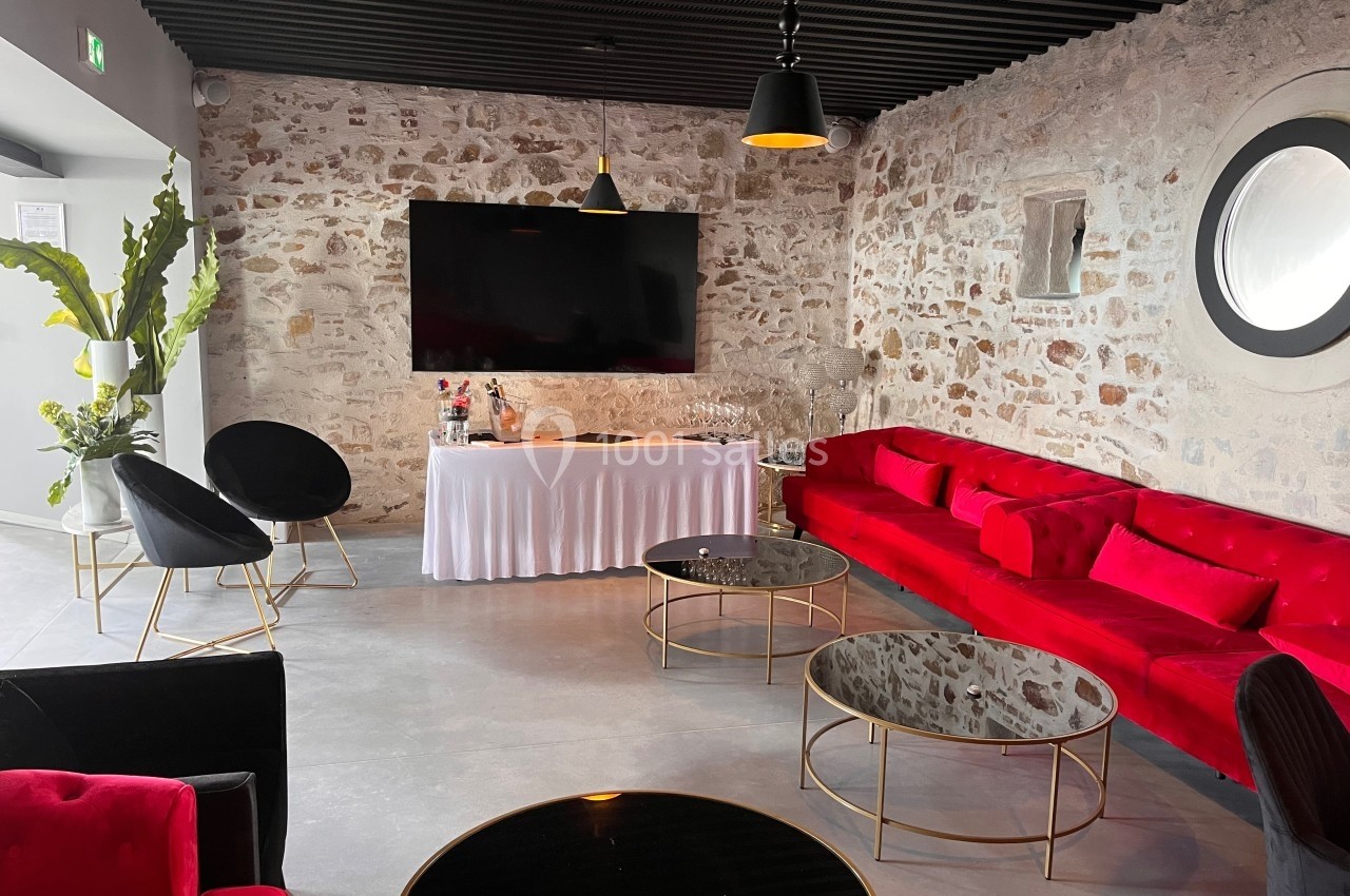 Salle moderne avec murs en pierre, canapé rouge, tables basses noires et écran mural, éclairée par des suspensions.