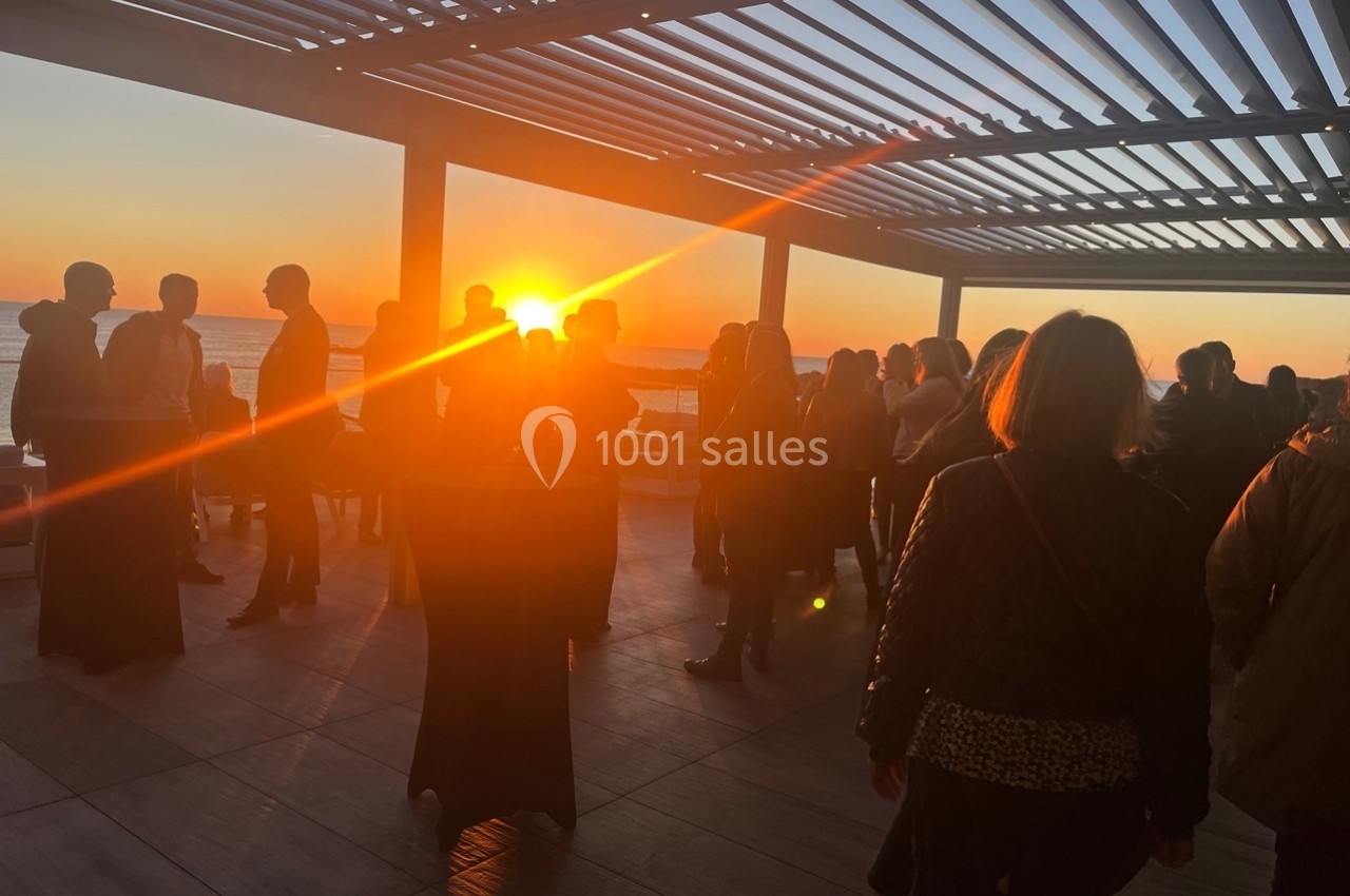 Groupe de personnes debout sur une terrasse au coucher du soleil, sous une pergola ouverte.