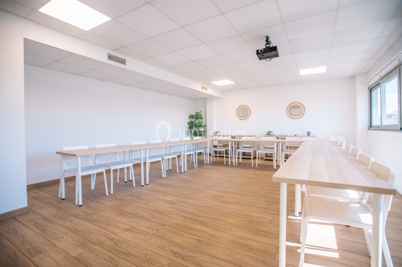 Salle de réunion lumineuse avec tables en bois clair disposées en U, chaises blanches et projecteur au plafond.