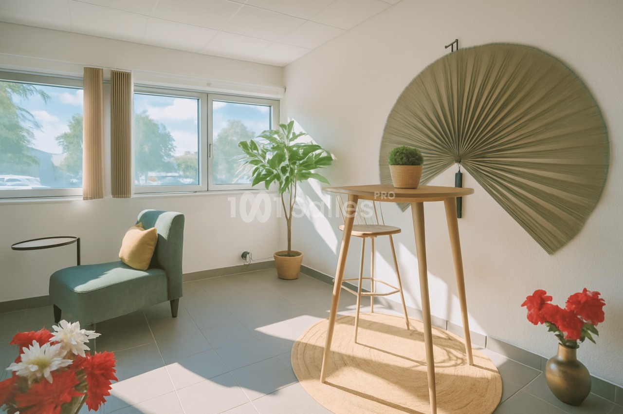Pièce lumineuse aux murs blancs avec un fauteuil vert d'eau, une table haute en bois entourée d'un tabouret, un grand éventail décoratif et des fleurs rouges au premier plan.