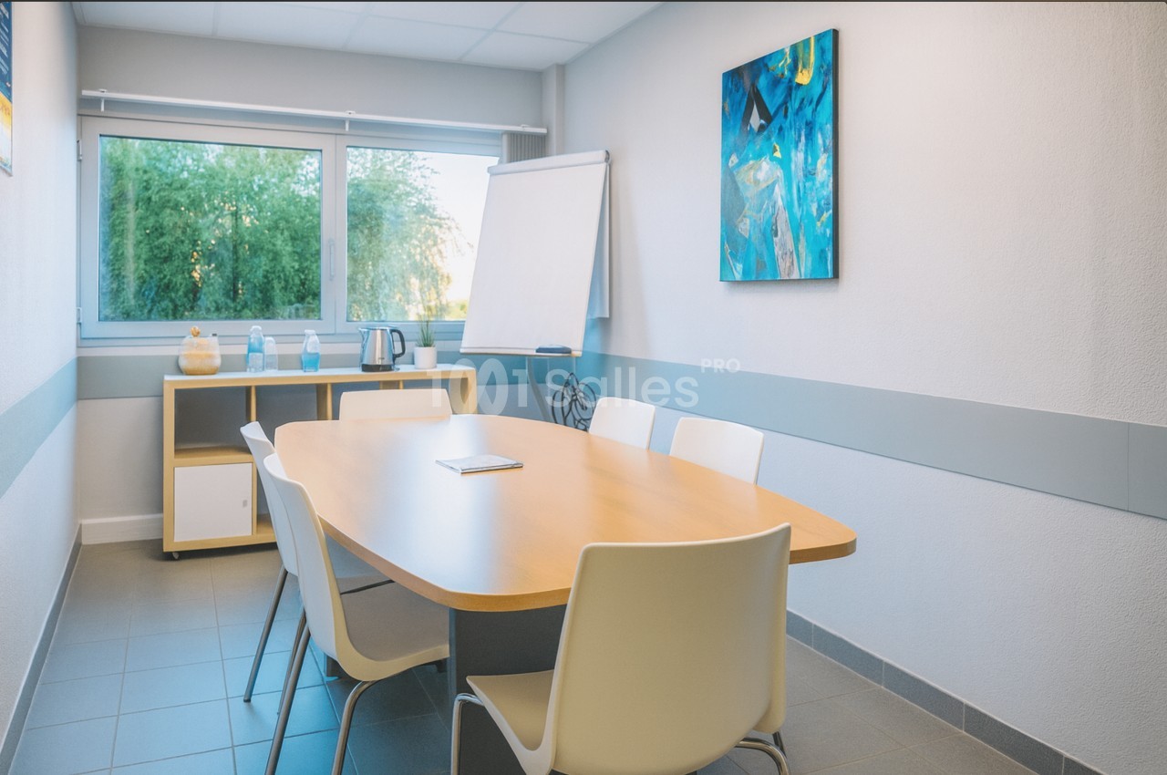 Petite salle de réunion avec une grande table ovale en bois entourée de chaises blanches, un tableau de conférence sur chevalet et une fenêtre donnant sur la verdure.