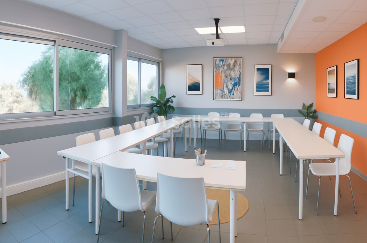 Salle de séminaire aux tables blanches disposées en U, chaises blanches, vidéoprojecteur au plafond et mur orange, avec grandes baies vitrées donnant sur des arbres.