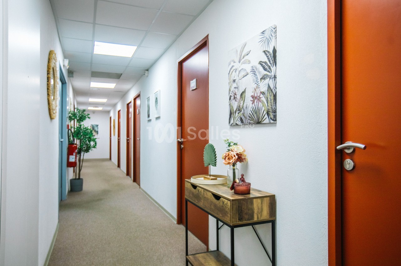 Couloir d'étage aux portes orange, moquette beige, tableau de feuillage tropical et console en bois avec fleurs au premier plan.