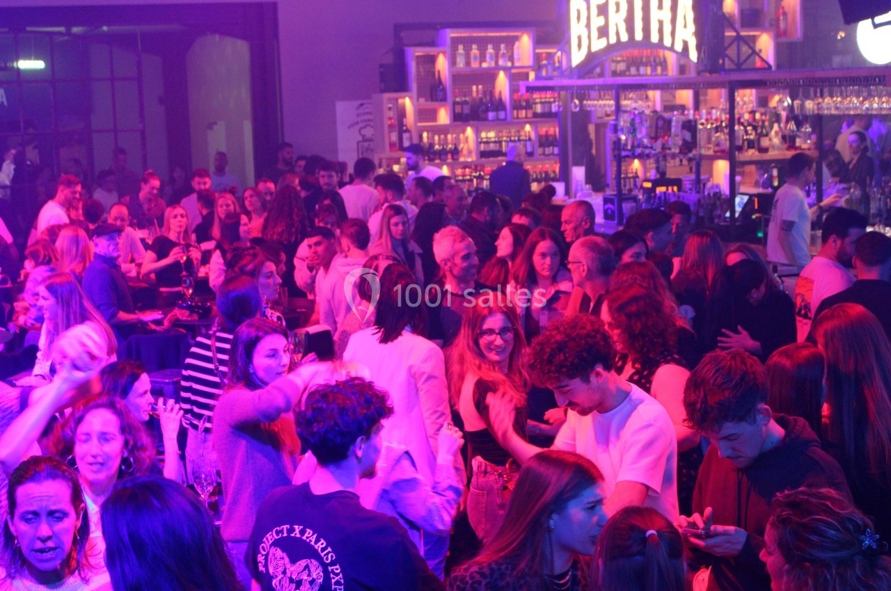 Foule dans un bar animé avec éclairage coloré, des personnes discutent et dansent devant un comptoir éclairé.