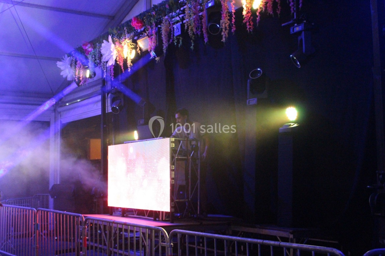 Un DJ mixe sur une scène éclairée par des projecteurs colorés, entourée de décorations florales suspendues.