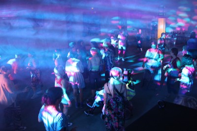 Personnes dansant et discutant lors d'une soirée en extérieur, éclairée par des lumières colorées et décorée de guirlandes.