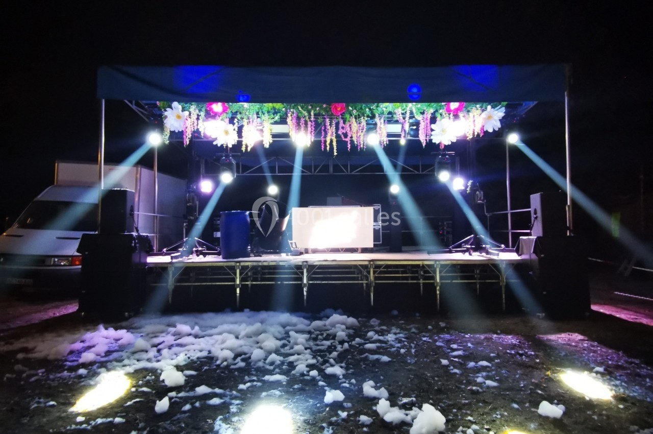 Scène extérieure éclairée la nuit avec des projecteurs, décorations suspendues et mousse au sol.